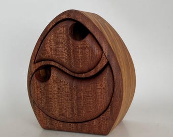 Op de afbeelding: Een druppelvormige houten doos met twee gebogen, in elkaar grijpende lades met ronde vingergrepen. De buitenkant toont een rijke, donkerbruine houtnerf op de voorzijde, gecontrasteerd door een lichtere, gladde beige houtnerf op het zijpaneel.
