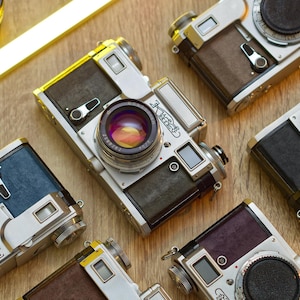 Puede incluir: Una colección de seis cámaras de película de 35 mm vintage plateadas y negras con cubiertas de cuero. Las cámaras tienen cubiertas de cuero de diferentes colores, incluyendo marrón, azul y morado.