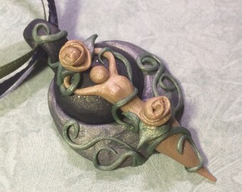 May include: A polymer clay pendant necklace featuring a dark grey and green base, a gold-coloured figure, and two rose-coloured flowers. The pendant is shaped like a crescent moon with vines and leaves.