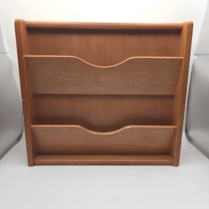 May include: A wooden desk organizer with three slots for storing documents or mail. The organizer is made of light brown wood and has a simple, functional design. The slots have curved openings for easy access.