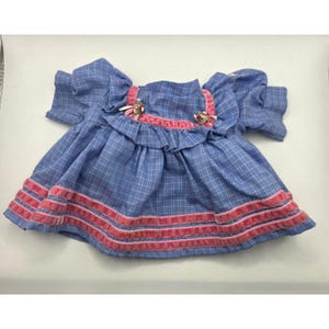 Pode incluir: Um vestido de boneca xadrez azul e branco com mangas curtas, corpete com babados e acabamento em fita de veludo rosa. A peça inclui dois pequenos laços decorativos na área do peito, com três fileiras horizontais de fita rosa na bainha.