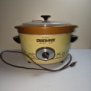 Może przedstawiać: W stylu vintage wolnowar Rival Crock-Pot w kolorze kremowym i brązowym. Urządzenie posiada szklaną pokrywę, czarne uchwyty oraz pokrętło sterowania z ustawieniami niskimi i wysokimi. Crock-Pot to serwer wyjmowany.