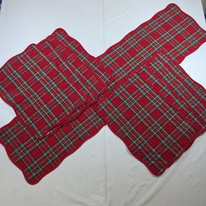May include: A set of quilted fabric placemats and a table runner featuring a red, green, and white plaid pattern. Each piece has a scalloped edge and a stitched diamond grid texture throughout the surface, creating a uniform look for table settings.