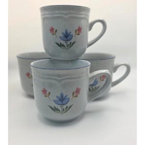 May include: Four ceramic mugs with a light gray speckled finish. Each mug features a blue floral design with pink accents and a blue rim. The mugs have curved handles and are stacked on top of each other.