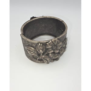 May include: Silver-toned metal cuff bracelet with a floral design. The bracelet features embossed flowers and leaves, creating a textured surface. The cuff is wide and has a hinged closure.