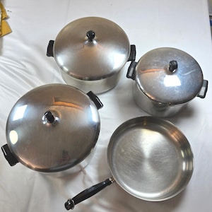 May include: A set of three silver-coloured metal pots with matching lids and one silver-coloured metal frying pan. Each pot features two black side handles and a black knob on the lid. The frying pan has a single long black handle extending from the side.