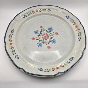 May include: A round, off-white ceramic plate with a blue rim and floral design. The center features a cluster of pink and blue flowers with green leaves. The rim is scalloped with additional floral accents in pink, blue, and brown.