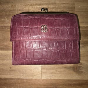 May include: A burgundy wallet with a crocodile skin texture. The wallet has a gold-colored clasp and a gold-colored emblem on the front. The wallet is closed and appears to be made of leather.