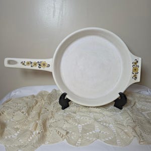 Pode incluir: Frigideira de cerâmica branca suja com uma longa alça e design floral em amarelo e marrom. A frigideira tem uma superfície de cozimento redonda e é suportada por um suporte preto. A alça e a lateral têm um padrão floral.