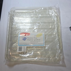 May include: A clear plastic Rubbermaid drainer tray encased in original packaging. The label reads Drainer Tray, Plateau-gouttoir, and lists dimensions of 36.3x38.9 cm. The rectangular tray features a series of raised parallel ridges.