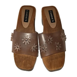 Pode incluir: Um par de sandálias com sola de madeira da marca Candie's. As sandálias têm uma base de madeira castanha, uma tira transparente adornada com flores transparentes e uma palmilha castanha com o nome da marca impresso.