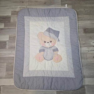 Pode incluir: Um edredom para bebé com um urso de peluche a usar um gorro de dormir e um pijama. O edredom tem um centro branco com a ilustração do urso, rodeado por uma borda xadrez azul e branca e uma borda de bolinhas azuis. O edredom é retangular.