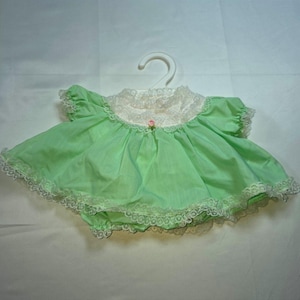 Könnte beinhalten: Ein hellgrünes Babykleid mit weißem Spitzenkragen und Spitzenbesatz. Das Kleid hat kurze Puffärmel, einen gerafften Rock und eine kleine rosa Rosenapplikation in der Mitte des Ausschnitts. Es wird auf einem weißen Kunststoffkleiderbügel präsentiert.