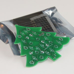 May include: A green Christmas tree-shaped circuit board with electronic components. The board has a network of lines and small components, with a silver package containing additional components in the background. A festive item for electronics enthusiasts.