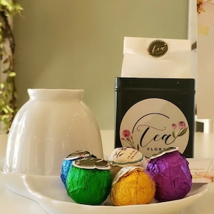 May include: A green tin with a white label that says "Tea Florals" and a white paper bag with a small sticker that says "Tea Florals". There are four flowering tea balls in a white bowl. The tea balls are wrapped in foil and are green, purple, yellow, and gold. A white sign with gold trim says "Flowering Tea Gift Set $9.25".