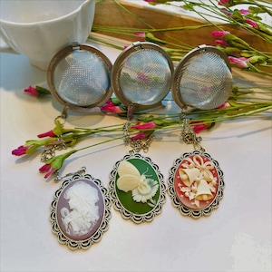 May include: Three stainless steel tea infusers with decorative cameo pendants. The pendants feature a woman's profile, a butterfly, and Christmas bells. The infusers are displayed with pink and green floral accents.