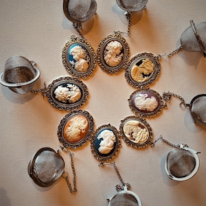 Può includere: Una collezione di infusori da tè con pendenti a cammeo in vari colori, tra cui blu, arancione e bianco. Ogni pendente è incastonato in una cornice ornata in stile antico e collegato a un infusore da tè in acciaio inossidabile con una catena.