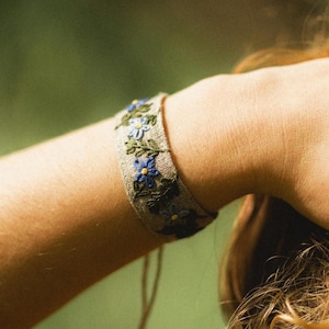 May include: A beige fabric bracelet with blue and green floral embroidery. The bracelet is wrapped around a person's wrist. The background is a blurred green, suggesting an outdoor setting. The bracelet is secured with a thin, beige string.