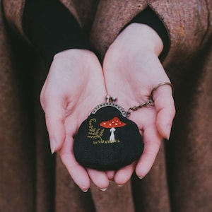 Pode incluir: Uma pequena bolsa preta com fecho de metal e chaveiro. A bolsa apresenta um desenho bordado de um cogumelo vermelho e branco com folhagem verde. O fundo está desfocado, sugerindo um ambiente ao ar livre.