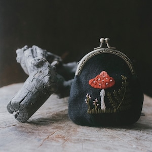 May include: A small, black coin purse with a metal clasp. The purse features an embroidered design of a red mushroom with white spots, green ferns, and small pink flowers. The purse is on a wooden surface.