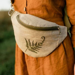 Pode incluir: Uma pochete bege com um desenho de bordado de samambaia. A bolsa tem um zíper verde escuro e um puxador de zíper prateado. A bolsa é usada sobre um vestido cor de ferrugem.