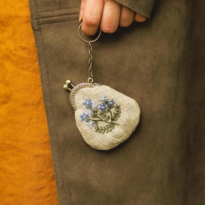 Peut inclure: Un petit porte-monnaie beige avec une broderie florale de fleurs bleues et de feuilles vertes. Le sac à main a un fermoir en métal et est attaché à un porte-clés. Le fond est un manteau marron et une chemise orange.