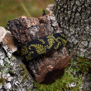 Könnte beinhalten: Ein schwarzes Stoffarmband mit olivgrünen, gestickten Blattmustern. Das Armband wird auf einem Stück strukturierter brauner Rinde präsentiert, umgeben von grünem Moos und einem verschwommenen natürlichen Hintergrund.