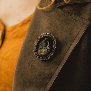 Op de afbeelding: Ovale broche met een zwarte achtergrond en een gedetailleerd bloemmotief in groen en goud. De broche is gezet in een decoratieve, antieke metalen lijst en is vastgemaakt aan een bruine jas.