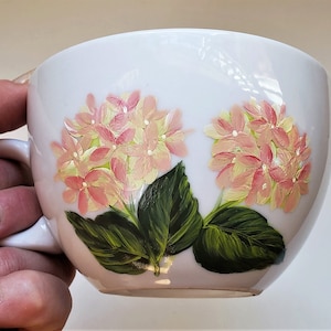 Può includere: Tazza da tè in ceramica bianca con un motivo floreale dipinto. Il disegno presenta fiori di ortensia rosa e gialli con foglie verdi. La tazza ha una forma curva e un manico.
