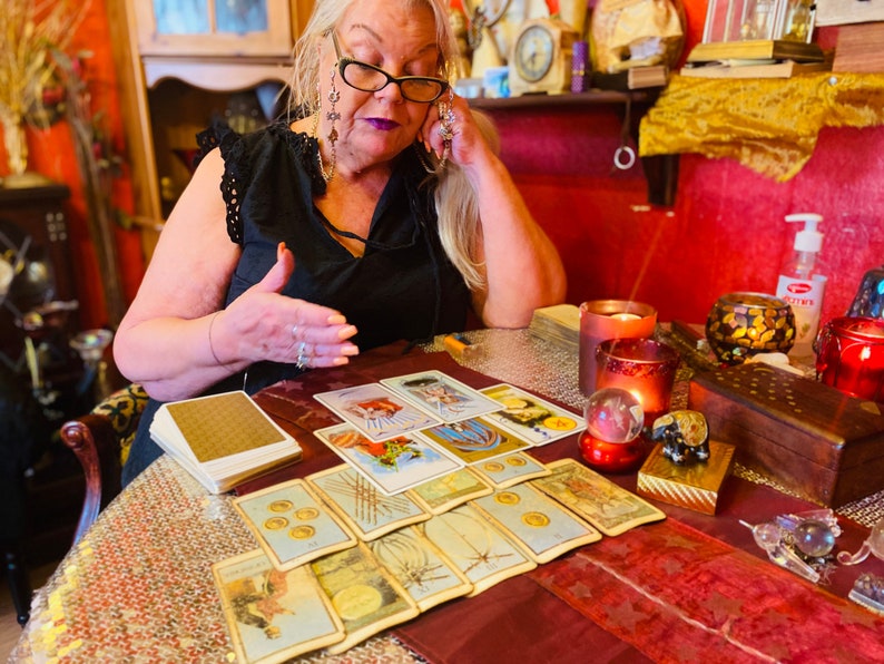 May include: A woman sits at a table with a spread of tarot cards, a crystal ball, and other divination tools. The table is covered in a red and gold tablecloth.
