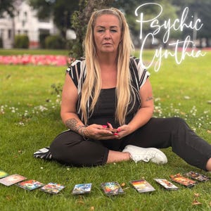 May include: A woman sits on grass with tarot cards laid out in front of her. She wears a black top and pants with a black and white striped top. The text "Psychic Cynthia" is written in cursive.