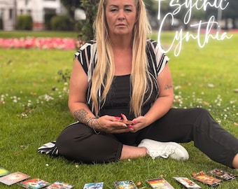 May include: A woman sits on grass with tarot cards laid out in front of her. She wears a black top and pants with a black and white striped top. The text "Psychic Cynthia" is written in cursive.