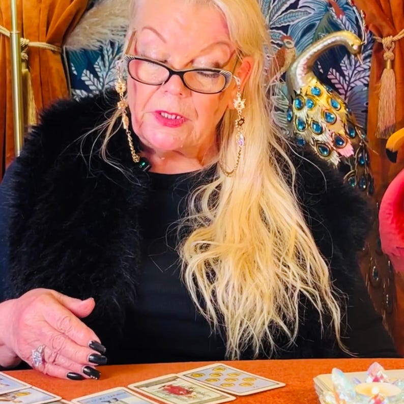 Puede incluir: Una mujer con cabello rubio y gafas est&aacute; sentada frente a una mesa con cartas de tarot extendidas. Lleva un cuello de piel negro y una camisa negra. Las cartas est&aacute;n extendidas en c&iacute;rculo, y hay un peque&ntilde;o cuenco blanco sobre la mesa.