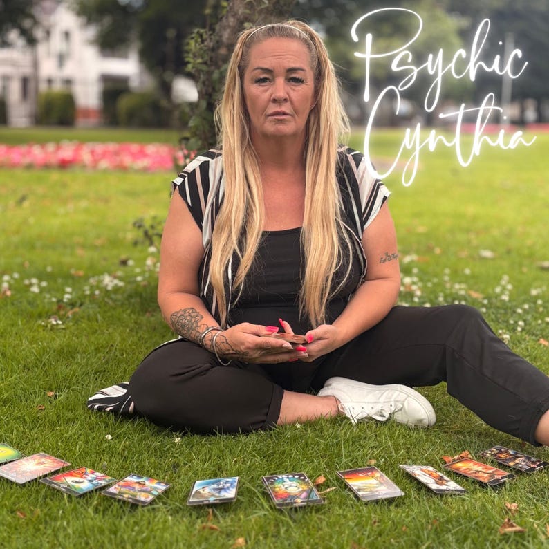 May include: A woman sits on grass with tarot cards laid out in front of her. She wears a black top and pants with a black and white striped top. The text "Psychic Cynthia" is written in cursive.