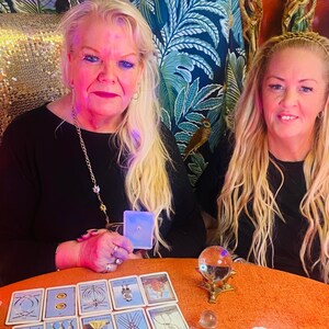 May include: Two women with long blonde hair are seated at a table with tarot cards and a crystal ball. One woman holds a card with a star on it. The table is covered in an orange cloth. A bird is perched on the curtain behind them.