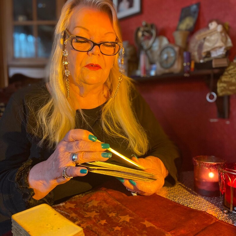 Puede incluir: Una mujer con el pelo largo y rubio est&aacute; sosteniendo una baraja de cartas de tarot y las est&aacute; barajando. Lleva una camisa negra y tiene las u&ntilde;as pintadas de un color turquesa brillante. Las cartas est&aacute;n en una pila y ella las est&aacute; sosteniendo con su mano izquierda. Su mano derecha est&aacute; sosteniendo un palo y lo est&aacute; moviendo a trav&eacute;s de la parte superior de las cartas. Hay una vela en el fondo.