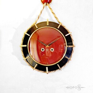 May include: A round, red-faced clock with black trim and gold accents. The clock is suspended from a white wall by a rope with a red knob at the top.