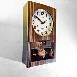 May include: A wooden wall clock with a white clock face and black hands. The clock face has black numerals and the brand name "LORUS". The clock has a lower compartment with decorative elements and the text "30 DAY".