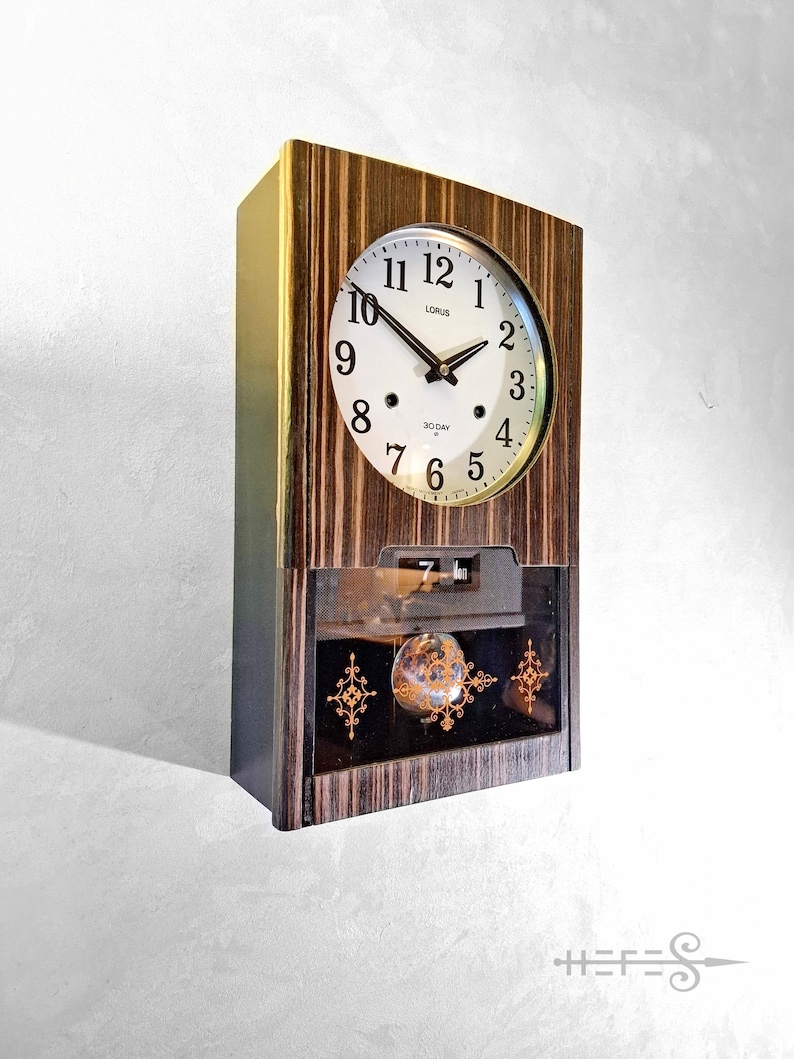 May include: A wooden wall clock with a white clock face and black hands. The clock face has black numerals and the brand name "LORUS". The clock has a lower compartment with decorative elements and the text "30 DAY".