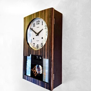 May include: A rectangular wooden wall clock with a round clock face. The clock face is white with black hands and numerals. The clock has a calendar display and a decorative glass panel. The clock is labelled "LORUS" and "30 DAY".