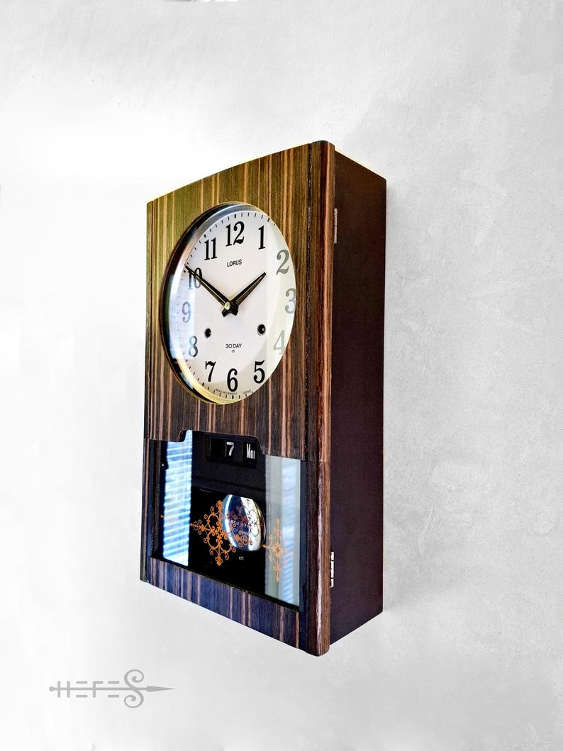 May include: A rectangular wooden wall clock with a round clock face. The clock face is white with black hands and numerals. The clock has a calendar display and a decorative glass panel. The clock is labelled "LORUS" and "30 DAY".