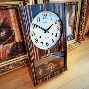 May include: A wooden clock with a striped design and a circular clock face. The clock face displays the brand "LORUS", black hands, and white numerals. The lower section has a glass panel with gold decorative patterns. The clock is positioned in front of framed pictures.