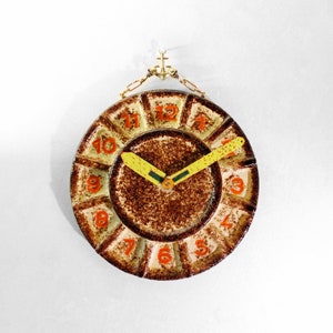 May include: A brown and yellow ceramic clock with orange numbers and a yellow hand. The clock is hanging from a gold anchor.