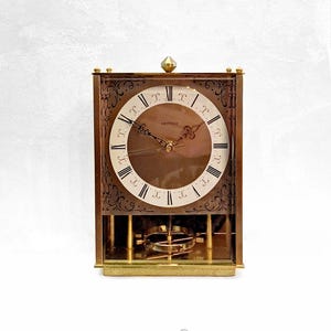 May include: A brass-toned, square-shaped clock with a white face and black Roman numeral markings. The clock has a pendulum and is made by the company "Hetich".