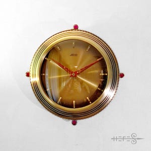 Può includere: Un orologio da parete vintage color oro con quadrante rotondo e lancette rosse. L'orologio ha una lunetta decorativa con tre gioielli rossi. Il quadrante dell'orologio ha una copertura in vetro a cupola. L'orologio è montato su una parete bianca.