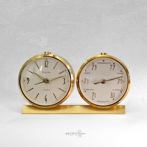 Puede incluir: Un juego vintage de reloj y barómetro en tono dorado. La esfera del reloj muestra la palabra "EUROPA" y el barómetro tiene las palabras "HEGEN", "STURMISCH", "VERANDERLICH", "SCHON" y "TROCKEN". El conjunto está montado sobre una base dorada.
