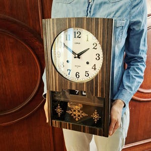 May include: A vintage Lorus 30-day clock in a wooden frame. The clock face is white with black numerals and hands. Below the clock face is a calendar display. The frame has a striped pattern.