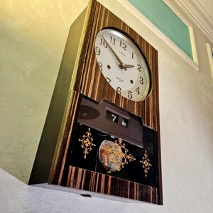 May include: A vintage wall clock with a wooden frame and a round clock face. The clock face is white with black numerals and hands. The clock also has a date display and decorative gold accents. The clock is mounted on a light-coloured wall.