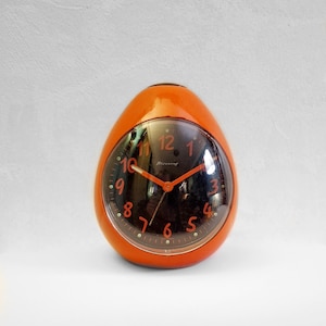 Può includere: Sveglia a forma di uovo arancione con quadrante nero e numeri bianchi. L'orologio ha un coperchio in vetro trasparente sul quadrante e dice "Blessing" sul quadrante.