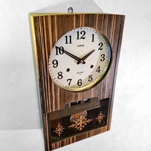 May include: A vintage Lorus wall clock with a wooden frame and a white clock face. The clock features black hands and numerals, with the text "LORUS" and "30 DAY" visible. The lower section has a glass panel with gold-coloured decorative elements.